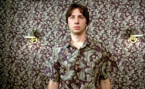 Zach Braff standing against a wall. His shirt and the wallpaper heavily blend together.